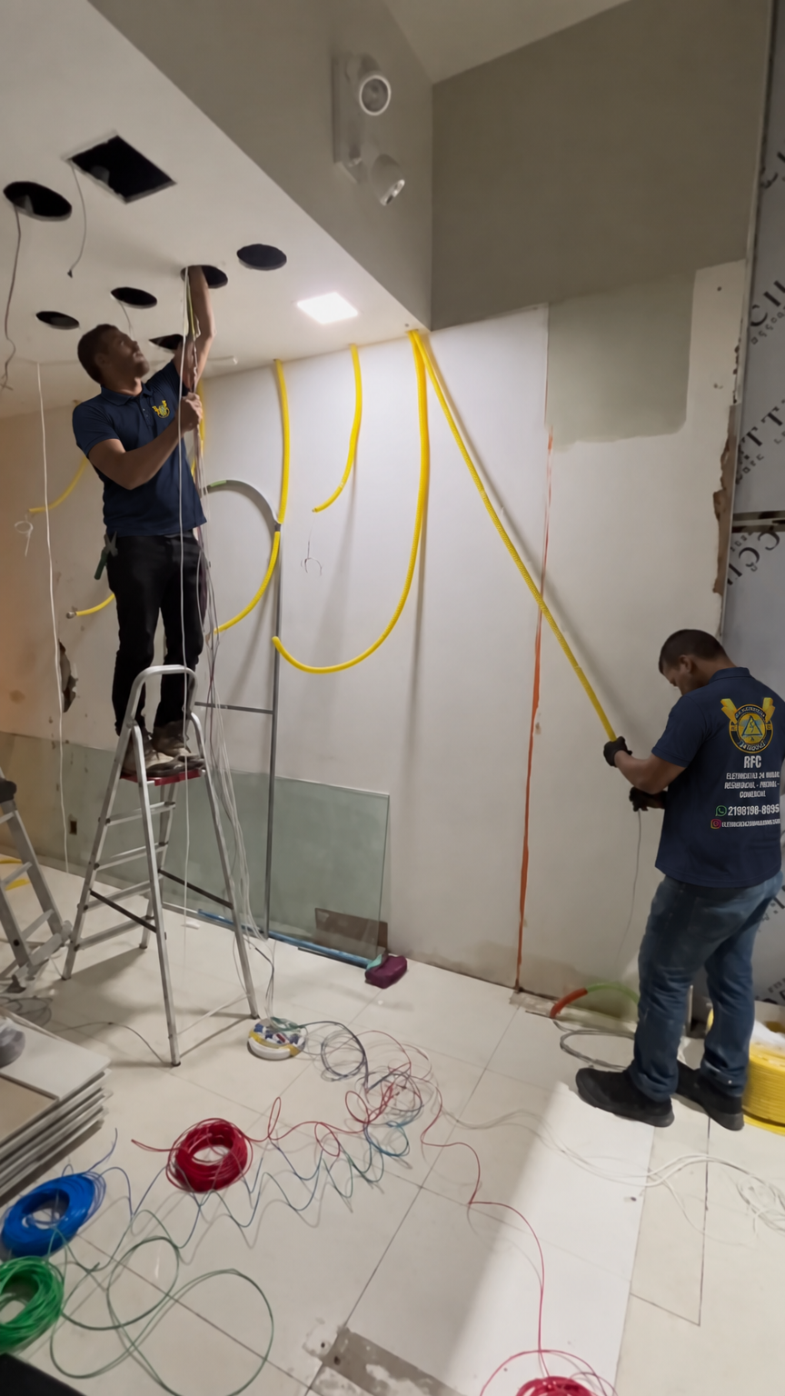 Dois profissionais passando fios pelo forro de gesso, com dutos amarelos na parede, com as camisas da RFC