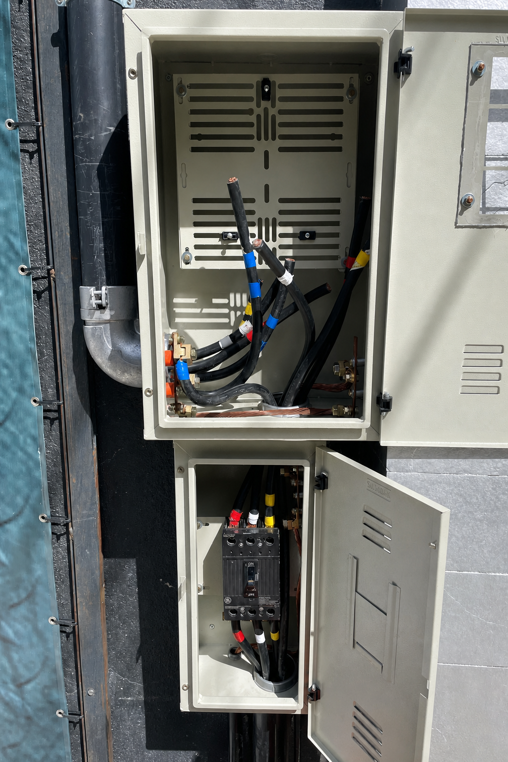 Quadro de entrada de energia com duas caixas metálicas sobrepostas, cabos identificados com fitas coloridas e disjuntor principal GE